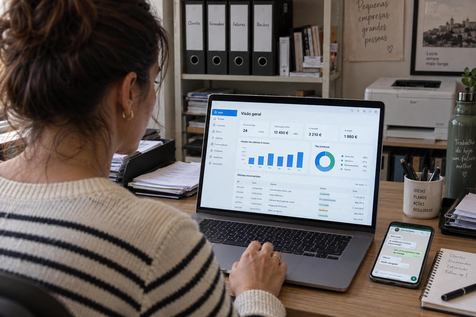 Dona de uma PME no back-office com o portátil a mostrar um ERP e o telemóvel com uma conversa de WhatsApp ao lado